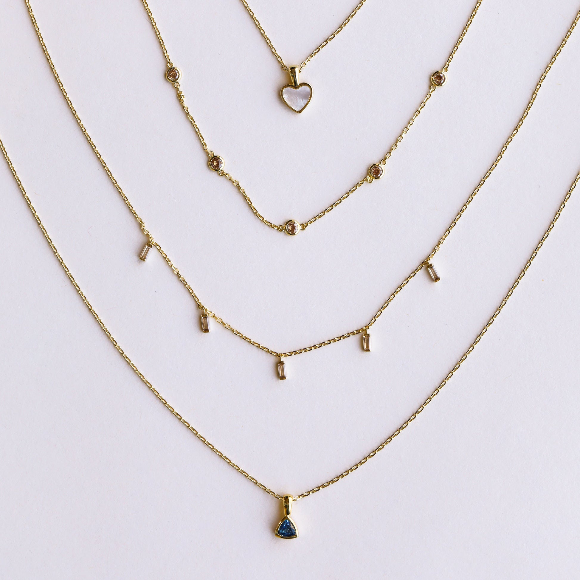 Four delicate gold necklaces by JaxKelly are arranged in a semi-circle. Discover the Mystery Necklace among heart, circles, bars, and blue triangle stone pendants—perfect for jewelry sales or as a surprise gift.