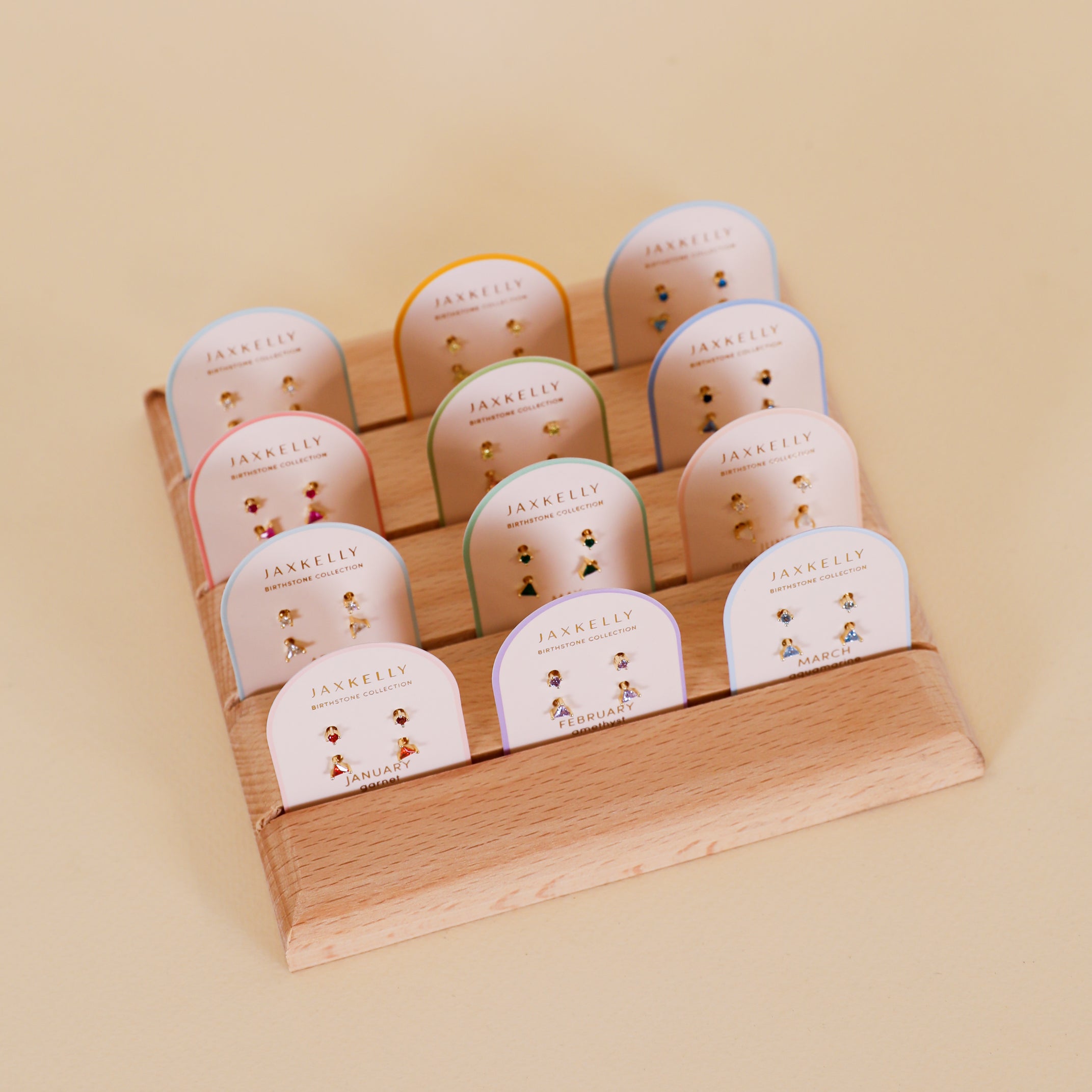 A wooden display stand holds twelve pairs of Birthstone Stud Earring Set - March, each on a white card, arranged in three rows against a light beige background.