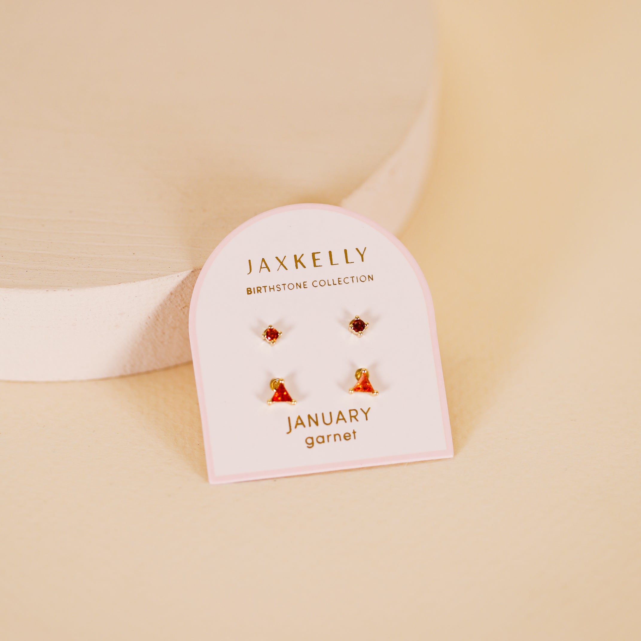 The Birthstone Stud Earring Set - January by JaxKelly, featuring gold-plated garnet studs, is shown on a light cream surface with a round white platform in the background.