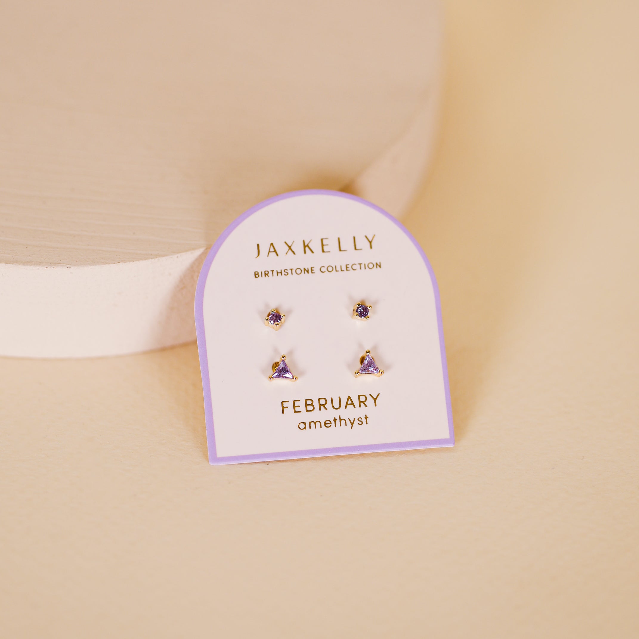 The Birthstone Stud Earring Set - February features amethyst studs from JaxKelly's Birthstone Collection, with sterling silver posts, displayed on a white and purple card against a soft beige background.