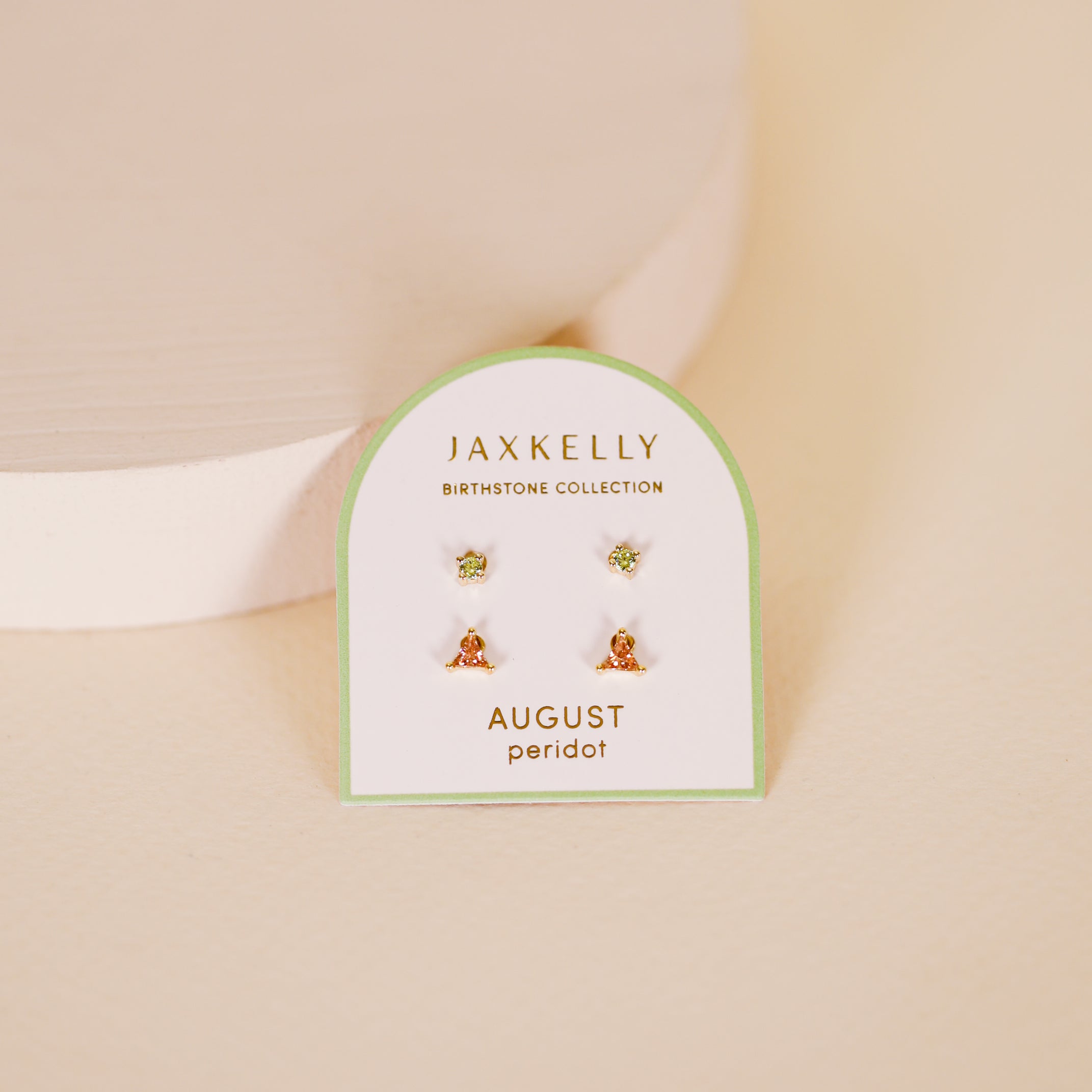 A pair of small birthstone stud earrings with light green stones, labeled "Birthstone Stud Earring Set - August," is displayed on a white card against a soft beige background.