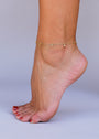 A close-up of a person’s foot with red-painted toenails, wearing the Solitaire Champagne Drop Anklet—an elegant gold-plated piece with small dangling charms—set against a plain light background.