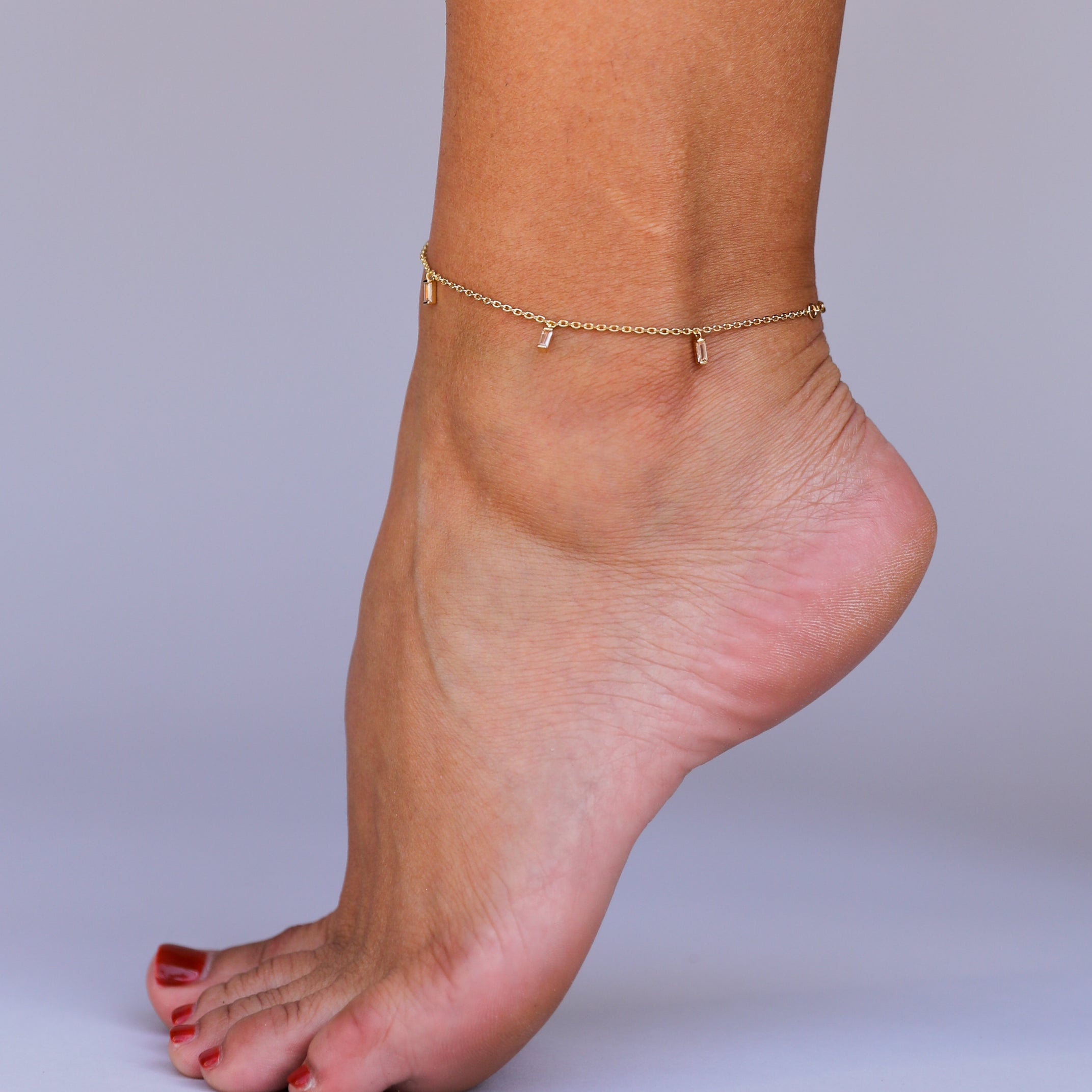 A close-up of a bare foot with red toenail polish wearing the Baguette Champagne Drop Anklet, an 18k gold plated piece adorned with small dangling charms, posed against a plain light background.