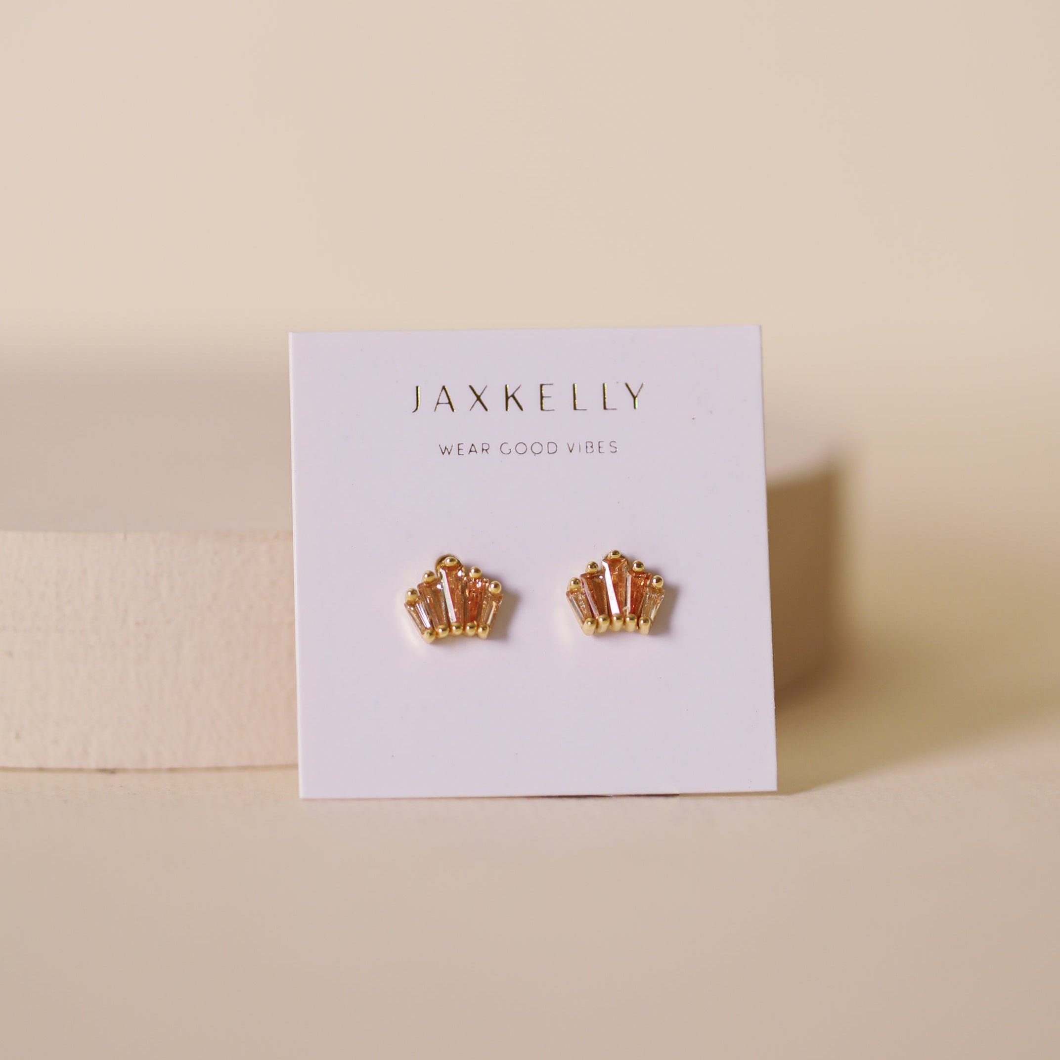 A pair of tarnish-free, 18K gold plated Champagne Baguette Crown Earrings by JaxKelly are shown on a white card labeled "Wear Good Vibes," against a neutral beige background.
