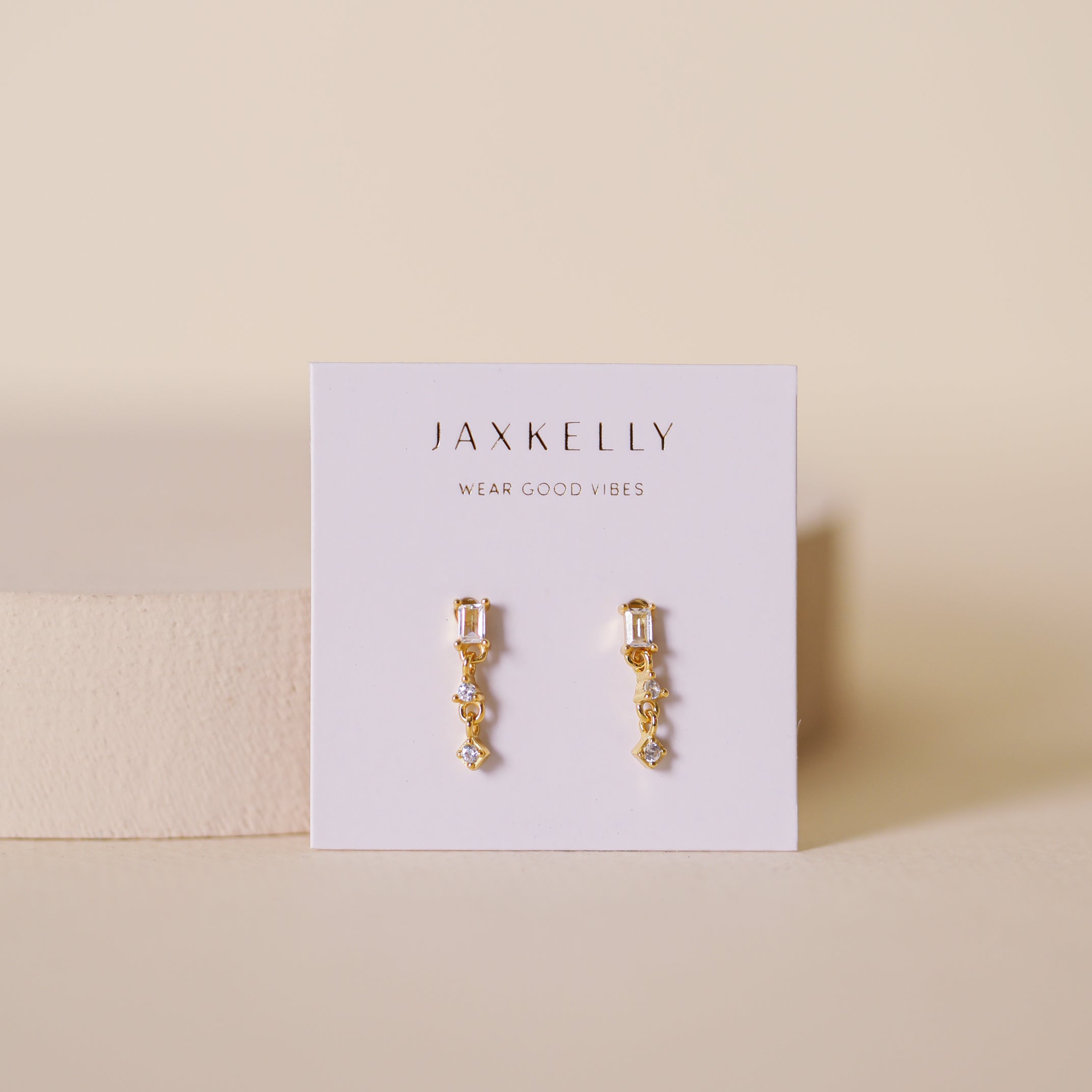 The Mini Baguette Drop - White earrings feature small geometric gold dangles with sparkling CZ stones and sterling silver posts, shown on a white JaxKelly Wear Good Vibes card against a softly lit beige background.