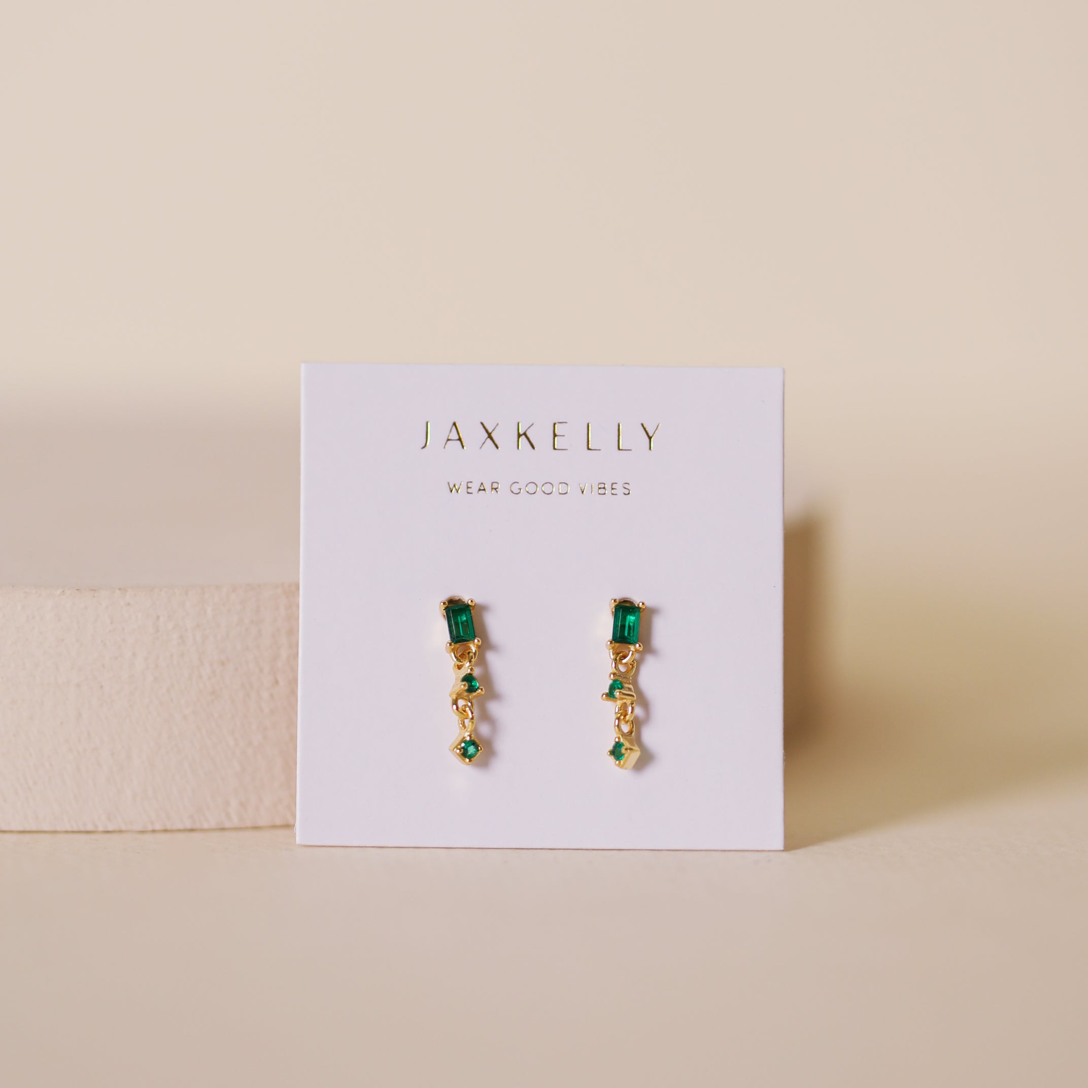 A pair of Mini Baguette Drop Earrings - Emerald, with sparkling CZ stones, shown on a white JaxKelly card reading WEAR GOOD VIBES, set against a neutral beige background.