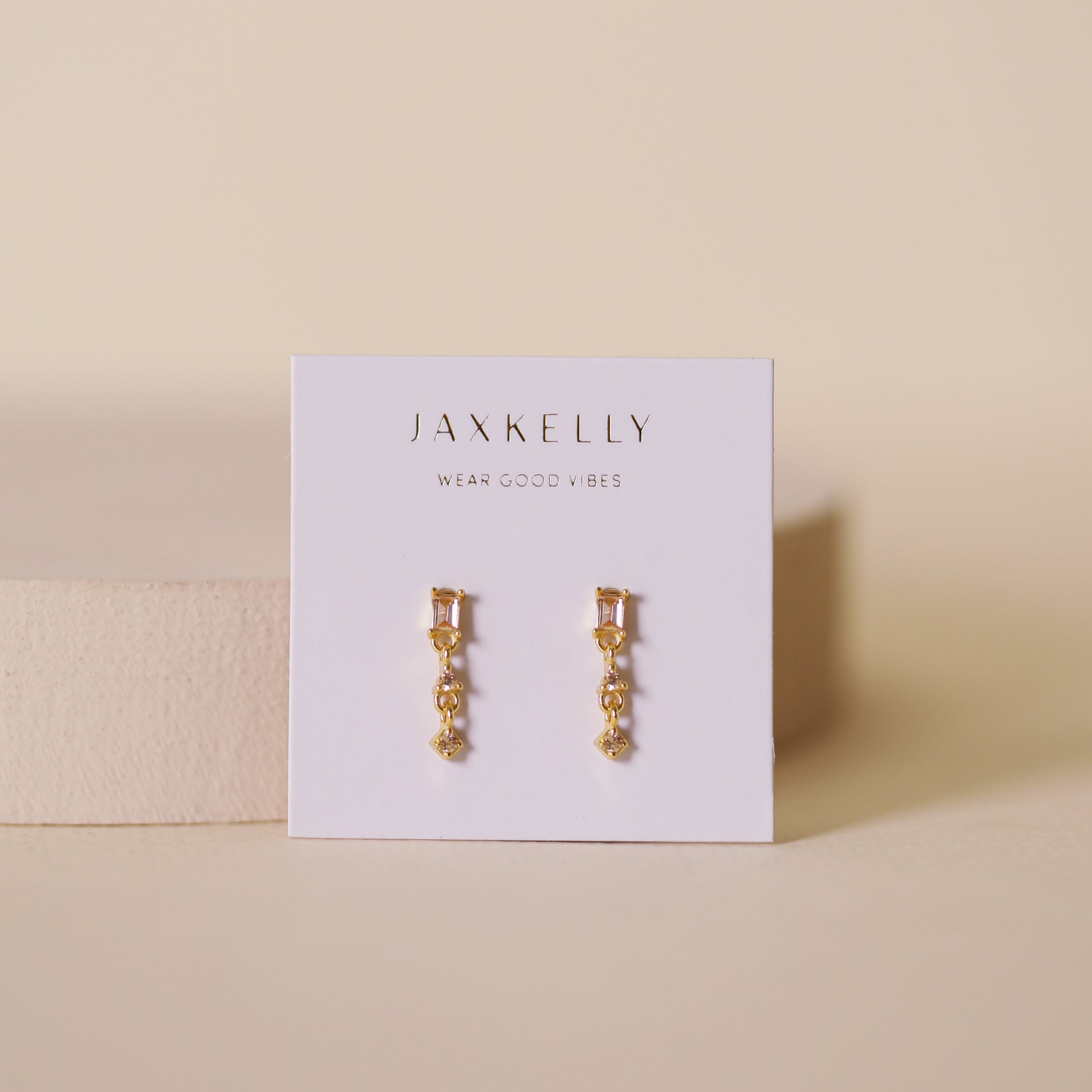 A pair of Mini Baguette Drop - Champagne gold plated earrings displayed on a white JaxKelly card reading "Wear Good Vibes," set against a neutral beige background.