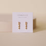 The JaxKelly Mini Baguette Drop - Champagne CZ earrings feature sparkling stones and delicate accents, displayed on a "Wear Good Vibes" card against a beige background—perfect gold drop earrings for anyone seeking tarnish-free style.