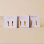 Three pairs of JaxKelly Mini Baguette Drop - White CZ earrings are displayed on white JaxKelly cards against a beige background. Each card features a unique tarnish-free design.