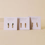 Three pairs of JaxKelly jewelry are displayed on branded cards against a beige background. The gold Mini Baguette Drop - Champagne CZ earrings feature champagne-colored stones for a timeless, tarnish-free look.