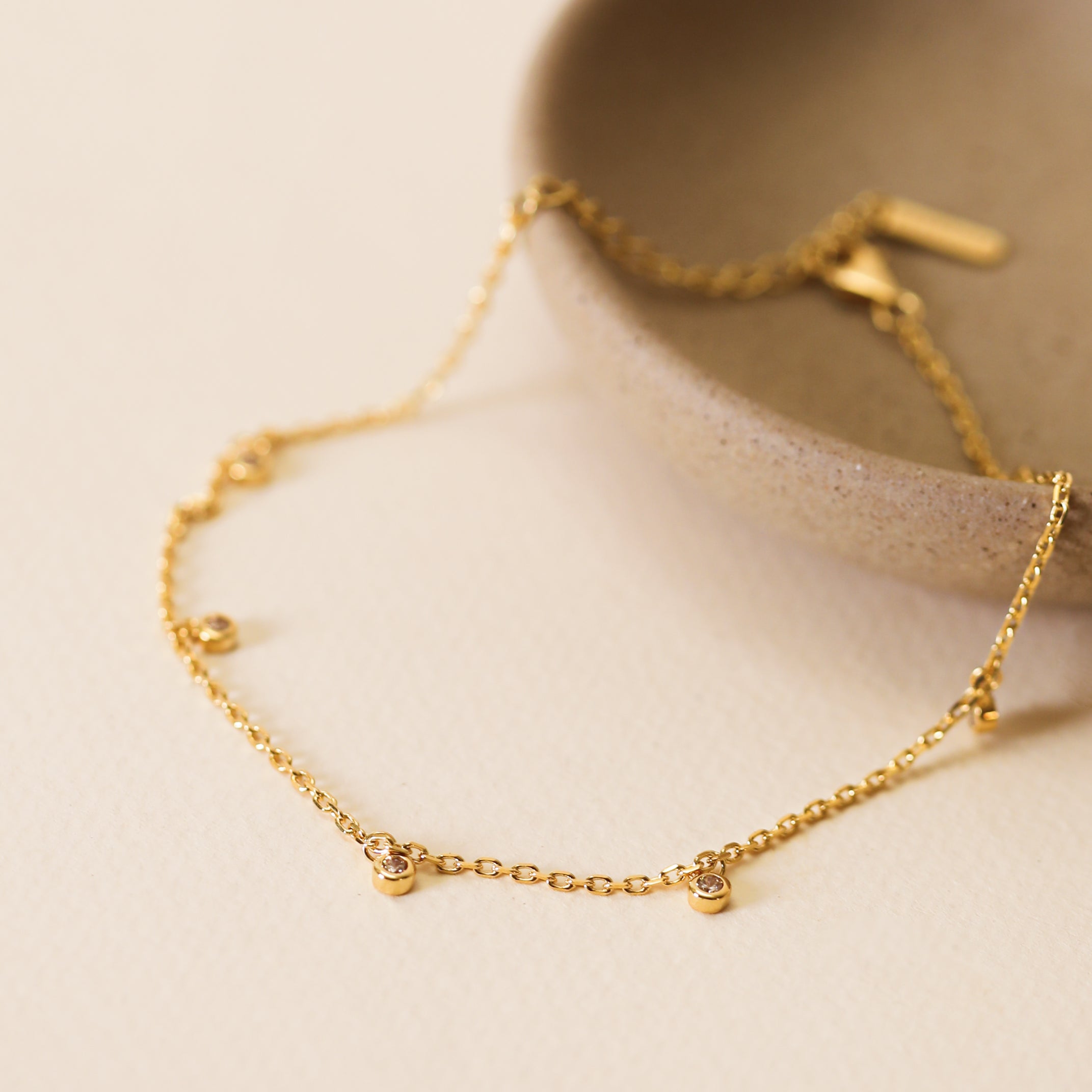 The Solitaire Champagne Drop Anklet, a delicate gold-plated piece with small gemstone accents, is displayed on a light surface and partially draped over the edge of a textured beige bowl.