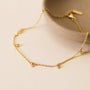 The Solitaire Champagne Drop Anklet, a delicate gold-plated piece with small gemstone accents, is displayed on a light surface and partially draped over the edge of a textured beige bowl.