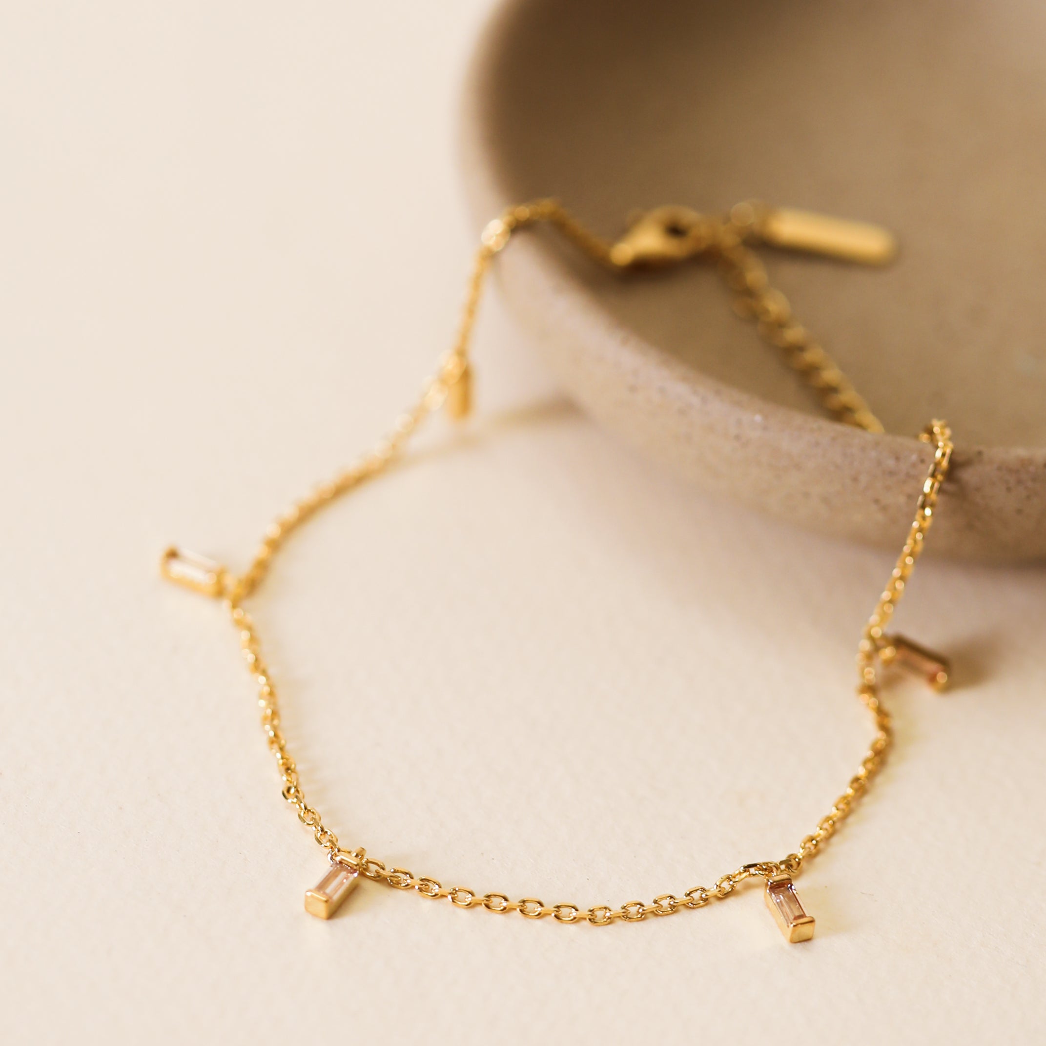 The Baguette Champagne Drop Anklet features a delicate 18k gold plated chain with rectangular charms and baguette CZ stones, showcased on a light beige surface beside a round, stone-colored dish.