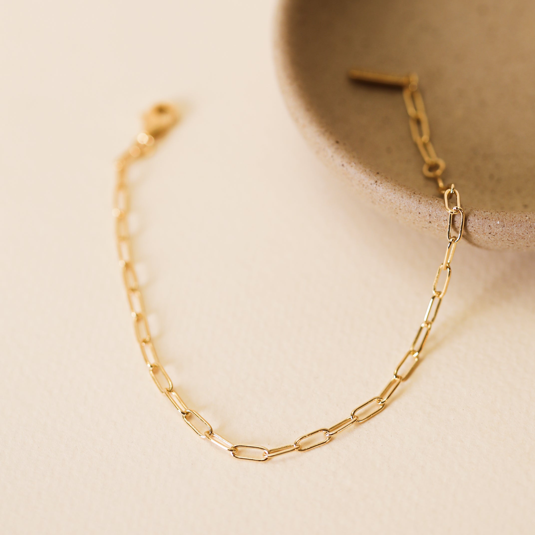 The Small Link Chain Bracelet, featuring delicate 18k gold-plated elongated links, is partially placed inside a round beige ceramic dish on a light textured surface.