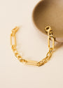 The Chunky Multi Link Chain Bracelet, featuring large rectangular links and plated in 18k gold, is displayed next to a round beige ceramic dish on a light textured surface.