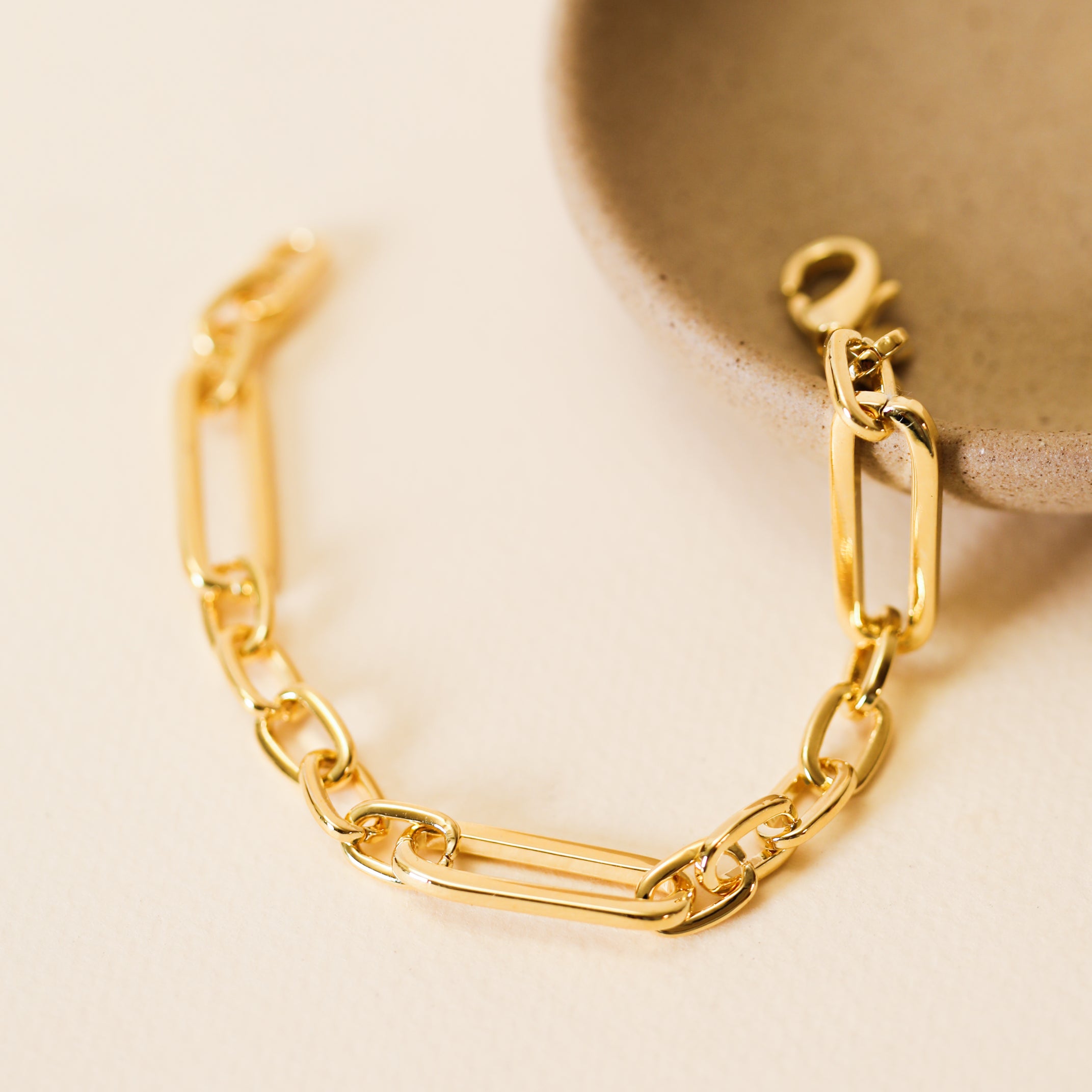 The Chunky Multi Link Chain Bracelet, featuring large rectangular links and plated in 18k gold, is displayed next to a round beige ceramic dish on a light textured surface.