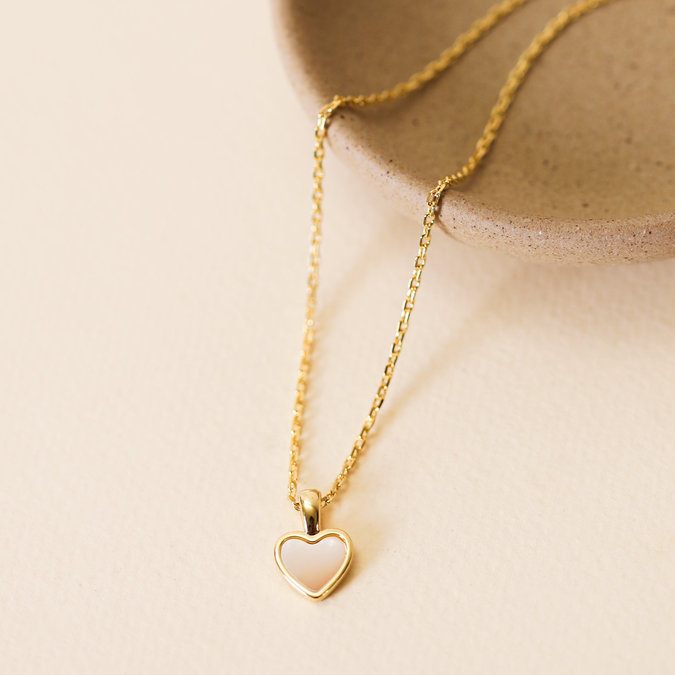 The Mother of Pearl Heart Pendant Necklace features a delicate heart-shaped pendant and is shown resting on a light beige surface beside a round ceramic dish.
