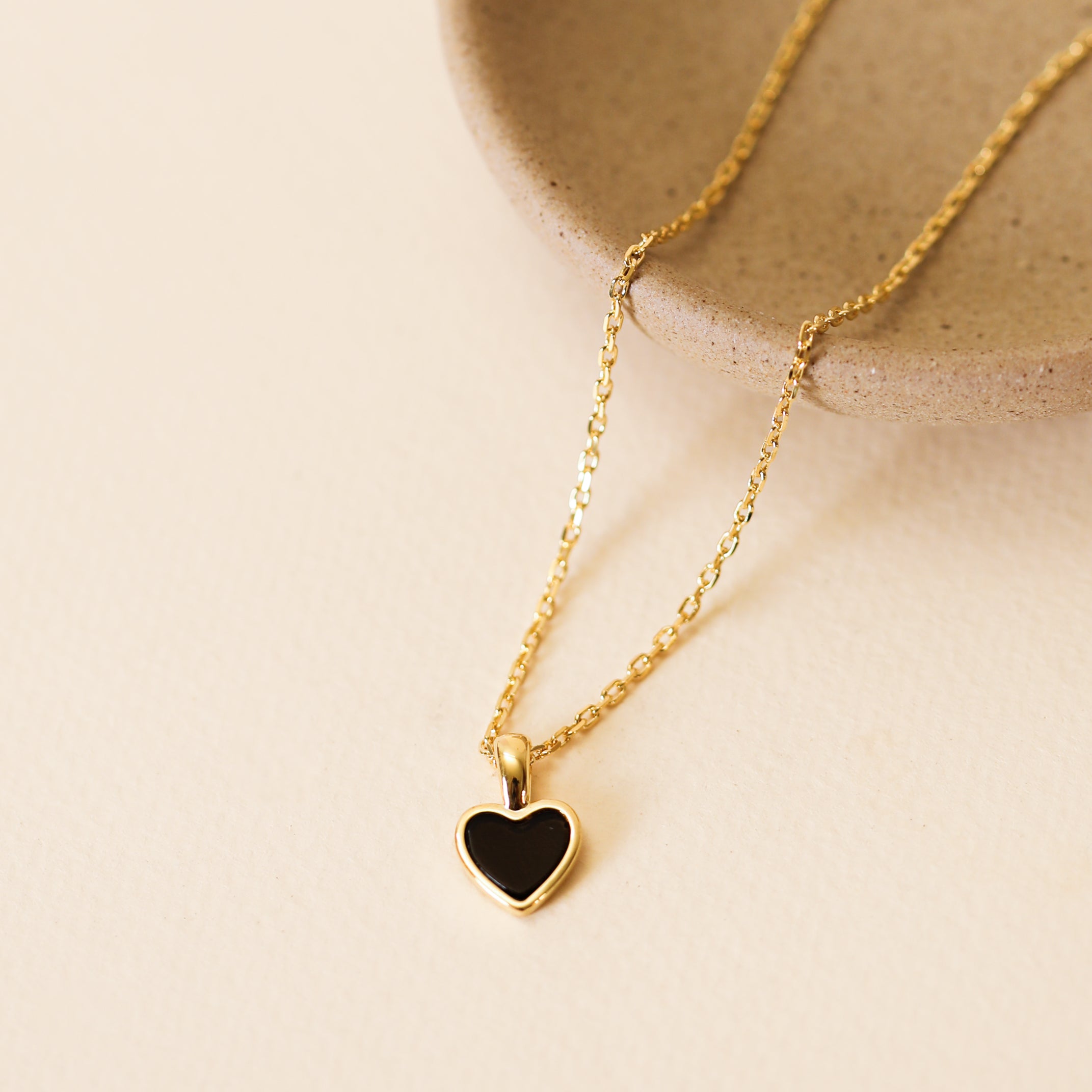 The Gemstone Heart Pendant Necklace - Black Tourmaline, featuring an 18k gold plated chain and a small heart-shaped pendant, is displayed on a textured beige surface beside a round ceramic dish.