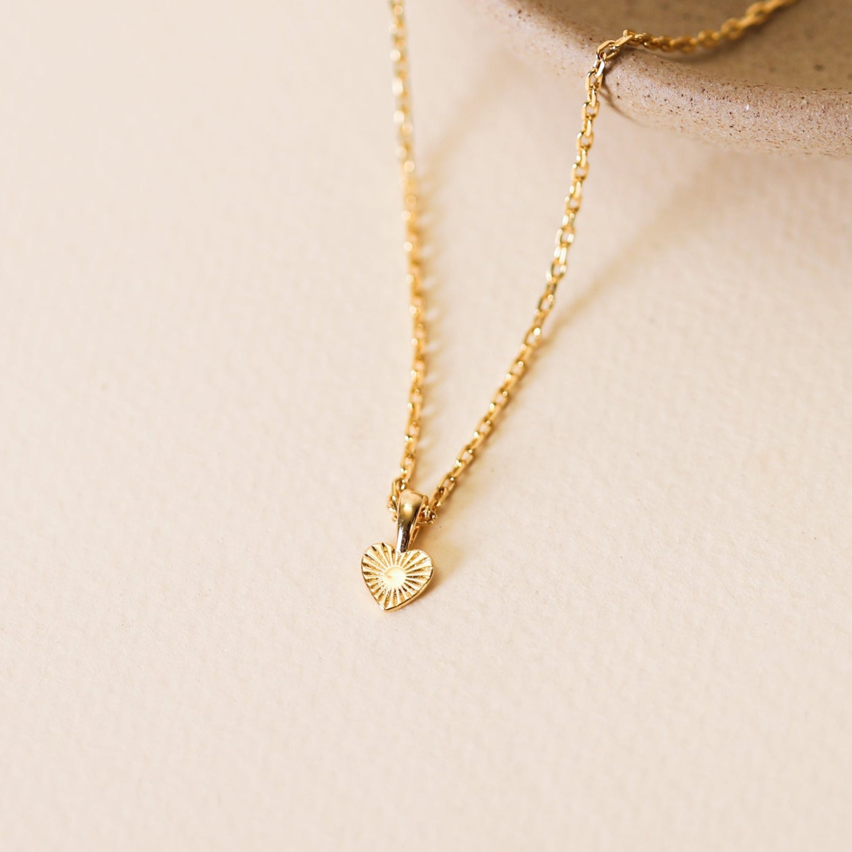 The Mini Heart Pendant Necklace, featuring a small textured gold heart charm and 18k gold plating, is displayed on the edge of a round beige ceramic dish against a light, neutral background.