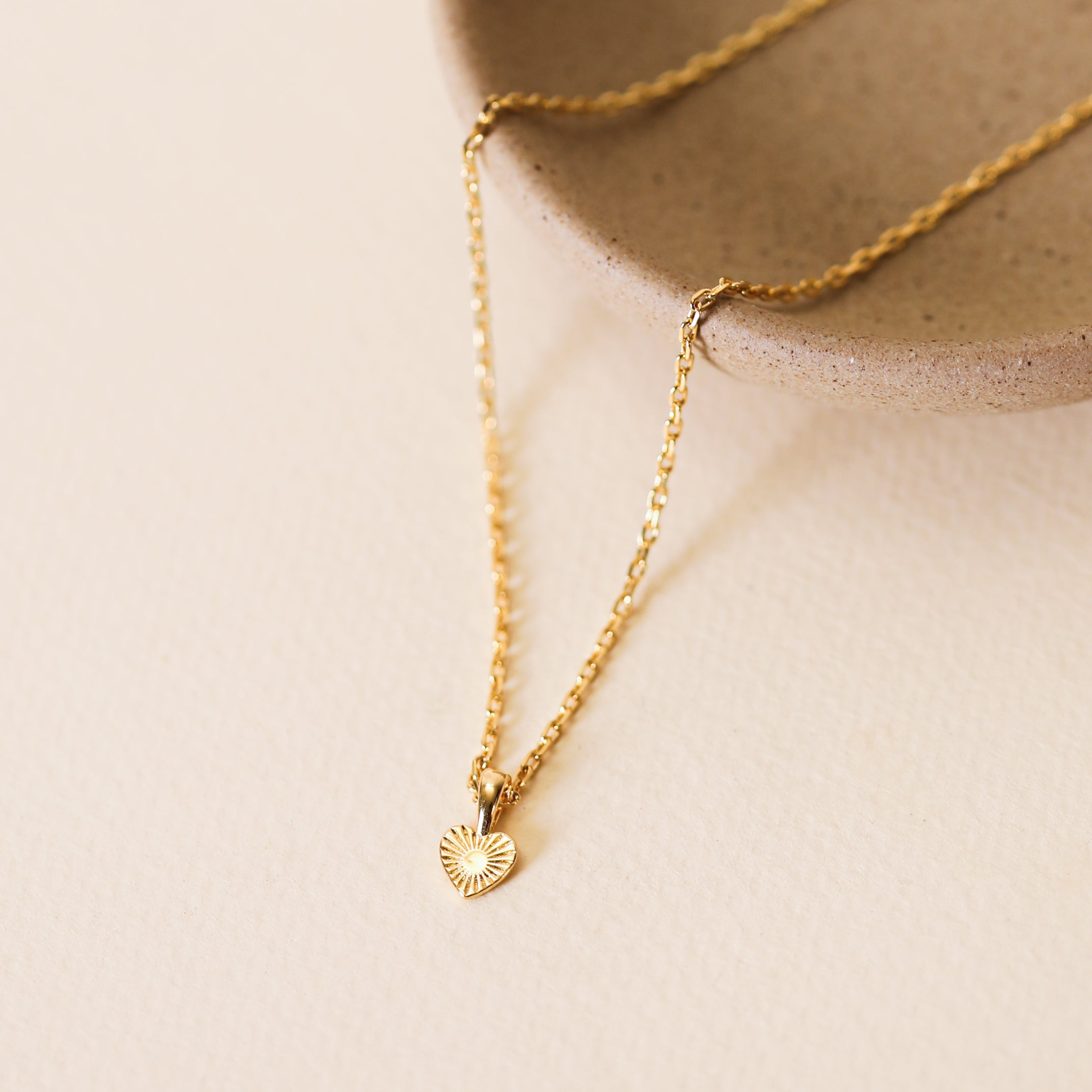 The Mini Heart Pendant Necklace, featuring a small textured gold heart charm and 18k gold plating, is displayed on the edge of a round beige ceramic dish against a light, neutral background.