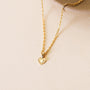 The Mini Heart Pendant Necklace, featuring a small textured gold heart charm and 18k gold plating, is displayed on the edge of a round beige ceramic dish against a light, neutral background.