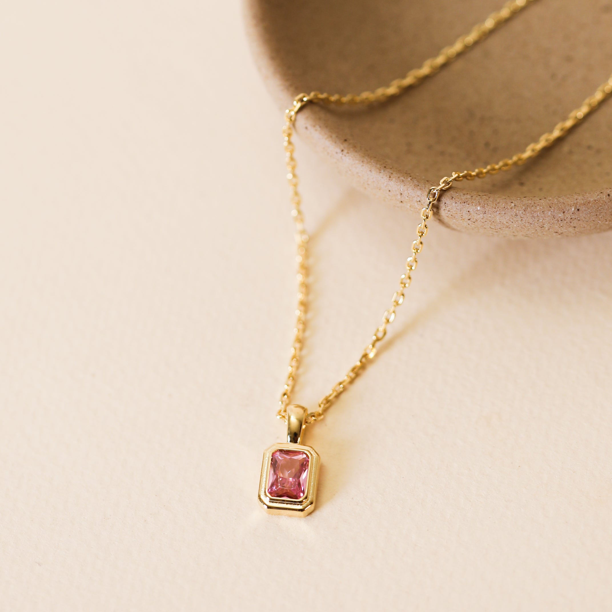 The Rectangular Baguette Pendant Necklace - Light Pink, a delicate 18k gold plated piece with a pink baguette stone pendant, is shown on a beige surface next to a round tan ceramic dish.