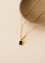 The Rectangular Baguette Pendant Necklace - Emerald Green showcases an 18k gold-plated chain and a green rectangular stone in a gold bezel, displayed on a light beige surface beside a round textured ceramic dish.