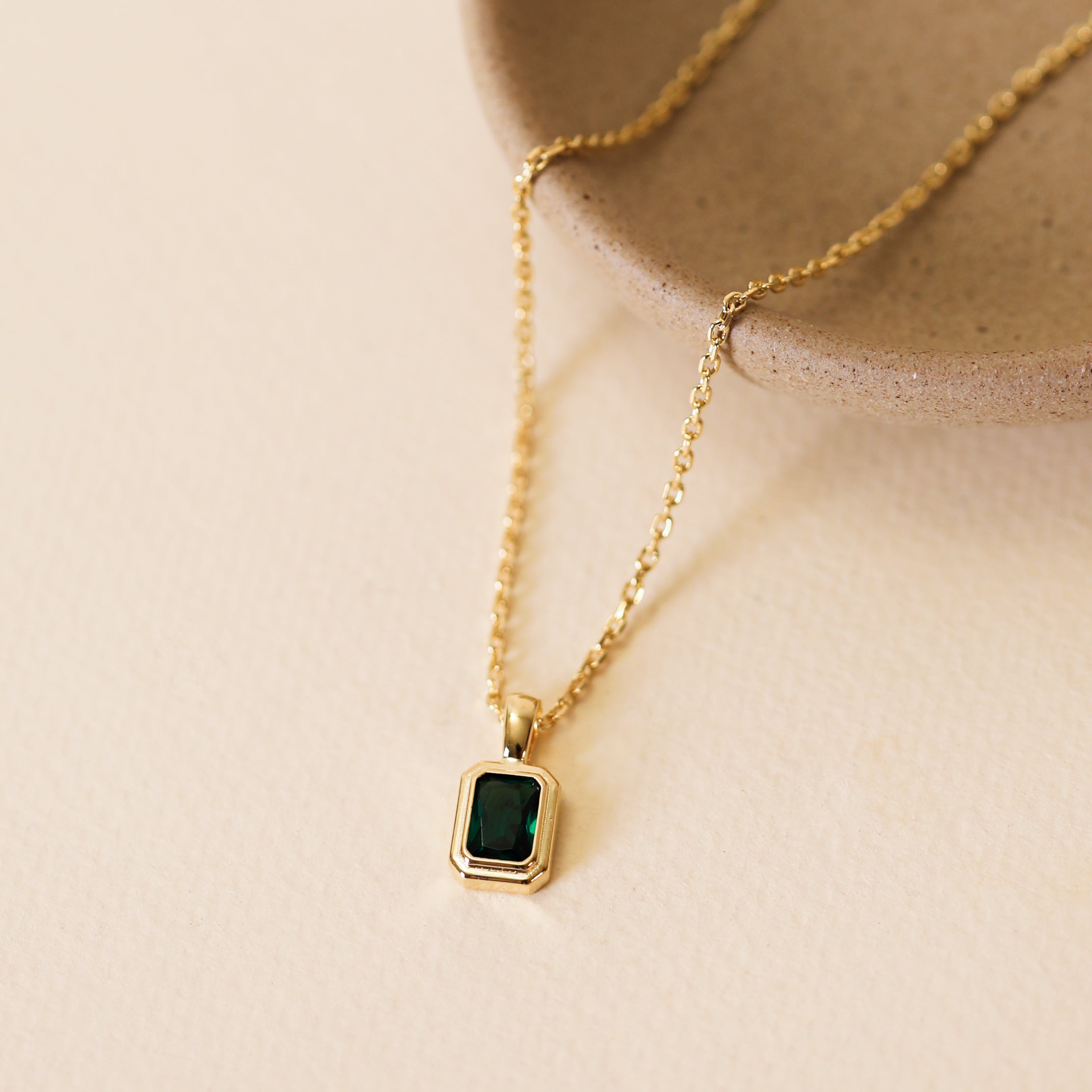 The Rectangular Baguette Pendant Necklace - Emerald Green showcases an 18k gold-plated chain and a green rectangular stone in a gold bezel, displayed on a light beige surface beside a round textured ceramic dish.