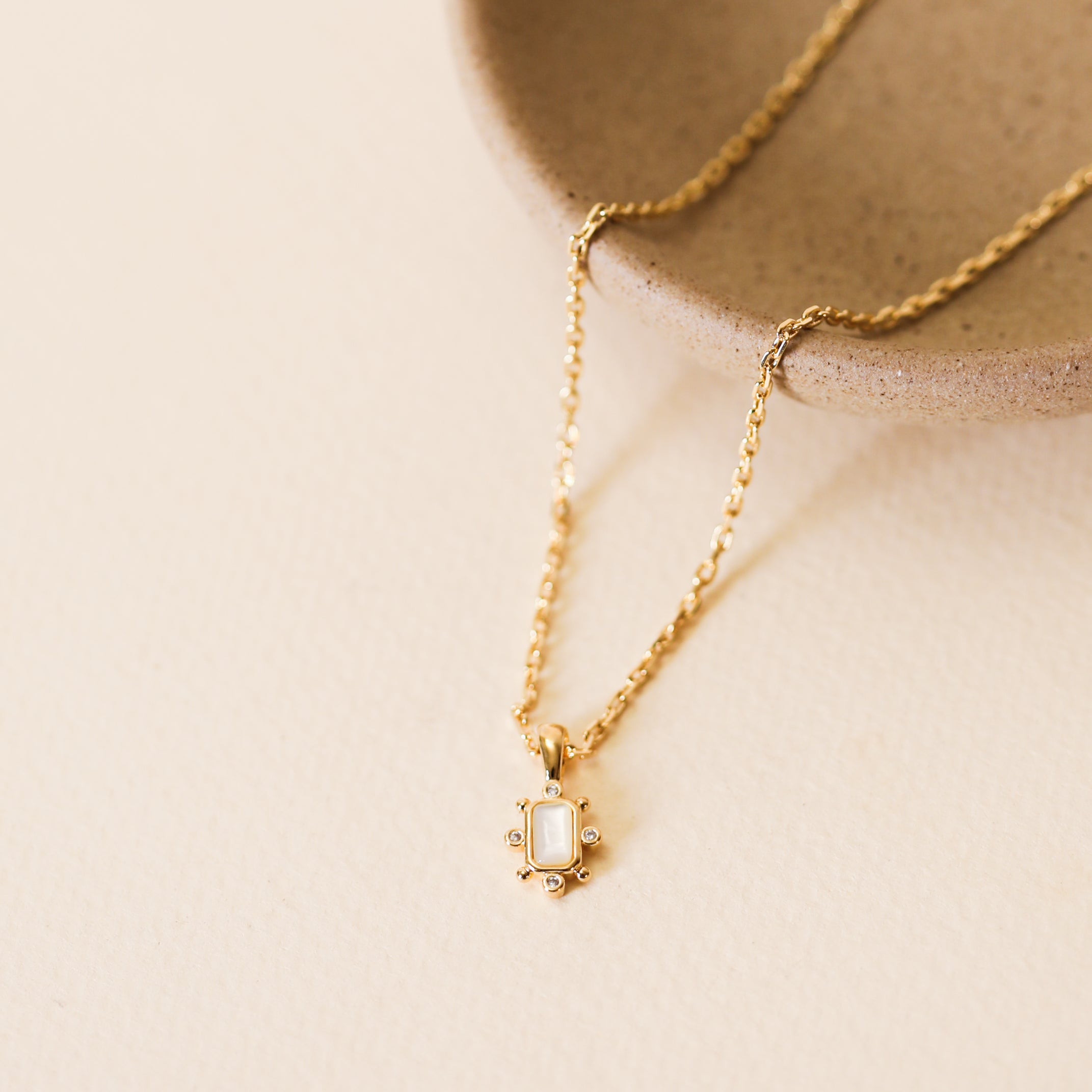 The Mother Of Pearl Gemstone Rectangular Pendant Necklace features an 18k gold plated chain, a small rectangular pendant with tiny decorative dots, and an adjustable clasp. Shown on a beige ceramic dish against a cream background.