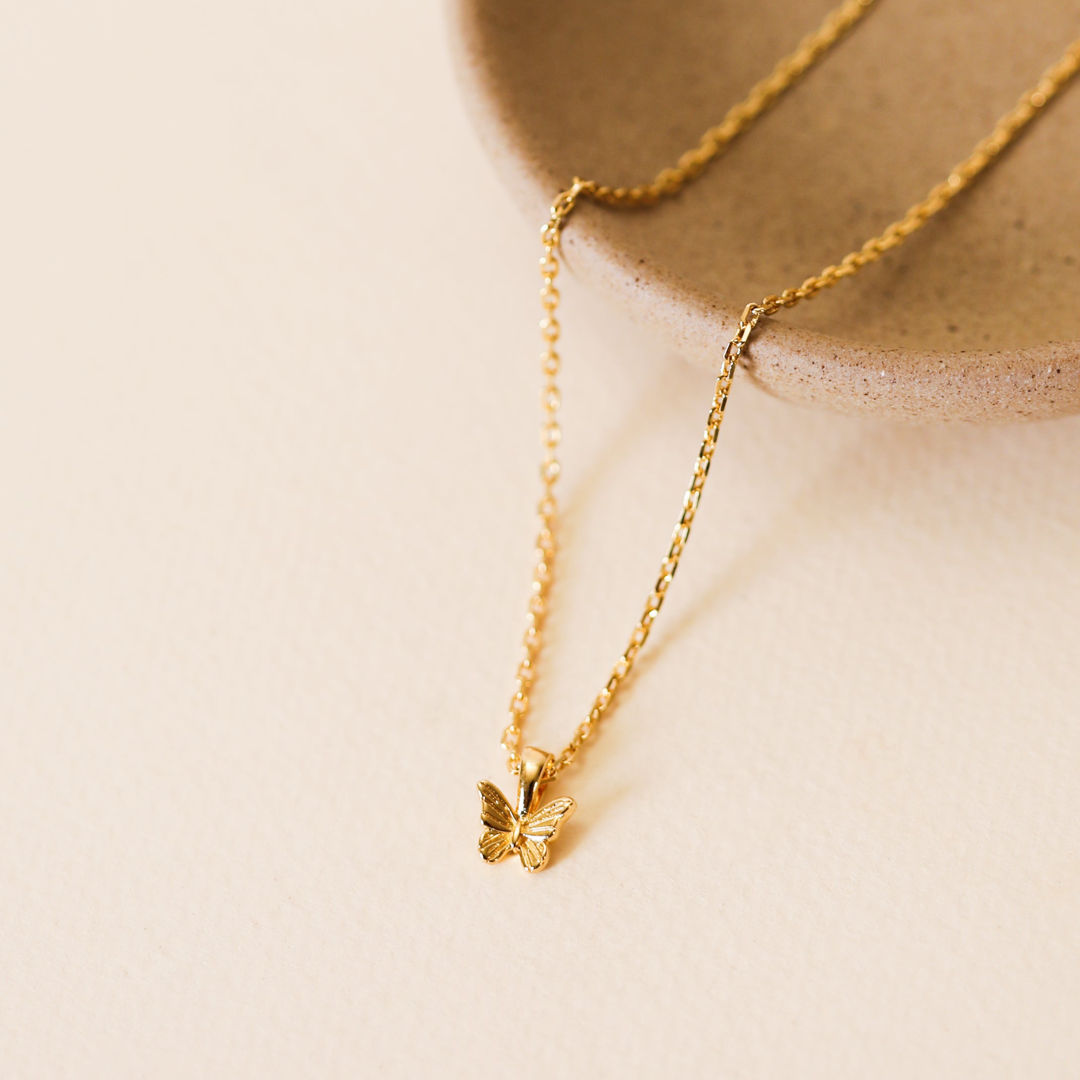The Mini Butterfly Pendant Necklace features a delicate gold butterfly on an 18k gold plated, diamond cut chain, displayed on a light textured surface beside a round beige ceramic dish.