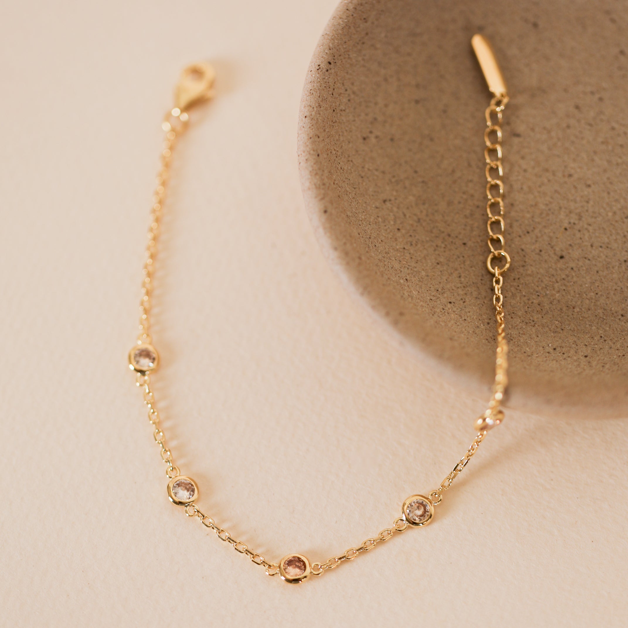 The Champagne Gemstone Bracelet features a delicate gold-plated chain with small round champagne stones, shown beside a round textured beige dish on a light cream background.