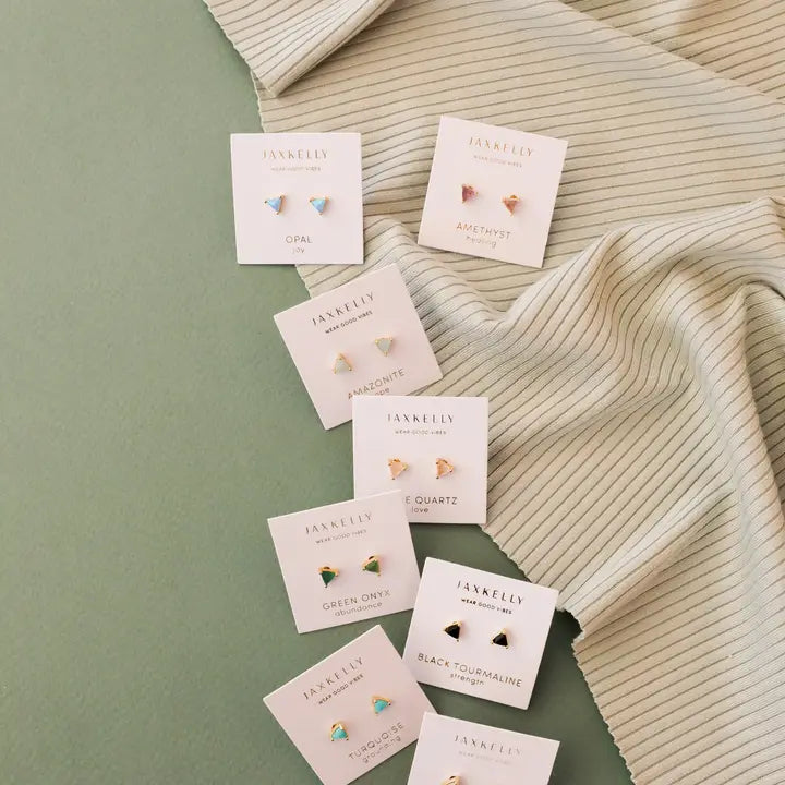 Seven pairs of JaxKelly Rose Quartz Mini Energy Gems - Love studs with 18k gold plating and sterling silver posts are displayed on branded cards over a soft green surface, beside cream ribbed fabric. Each card features a rose quartz gem and the JaxKelly name.