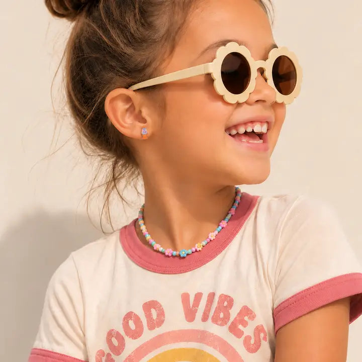 Smiling young girl wearing round, flower-shaped sunglasses, a Good Vibes t-shirt, a colorful beaded necklace, and JaxKelly Lava Lamp Enamel Flat Back Kids Earrings for sensitive ears, looking to the side against a light background.
