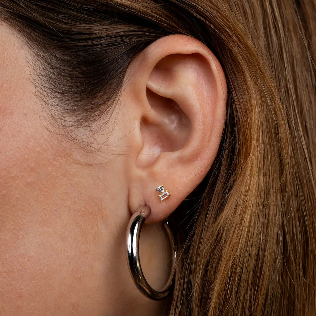 Close-up of a person’s ear wearing JaxKelly’s Double Stud Stack - White CZ: an 18k gold plated, white CZ baguette geometric stud in the upper lobe and a bold gold hoop in the lower lobe. Both have a durable protective coating. Light brown hair visible.