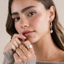 A young woman with long brown hair wears JaxKelly’s Twisted Pearl Drop Hoop - Gold Earrings, paired with gold rings and a delicate gold necklace. She has natural makeup and gently touches her face while looking into the camera.