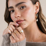 A woman with light brown hair and natural makeup wears JaxKelly’s Floating Gemstone Ring – White Opal, gold jewelry, pearl hoop earrings, and a necklace. She touches her chin and lips while wearing a beige sweater.