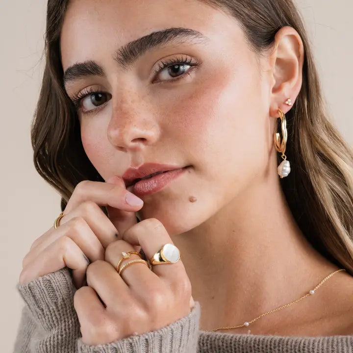 A woman with wavy brown hair poses in natural makeup and a brown sweater, wearing JaxKelly's Trio Cluster Gold Earrings – Pearl, plus stacked rings and a delicate necklace. Her hand is near her lips.