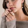 A woman with light skin and brown hair wears JaxKelly's Dainty Pearl Station Necklace, gold rings, and hoop earrings. Thoughtful in a beige knit sweater, she gently touches her lips and chin.