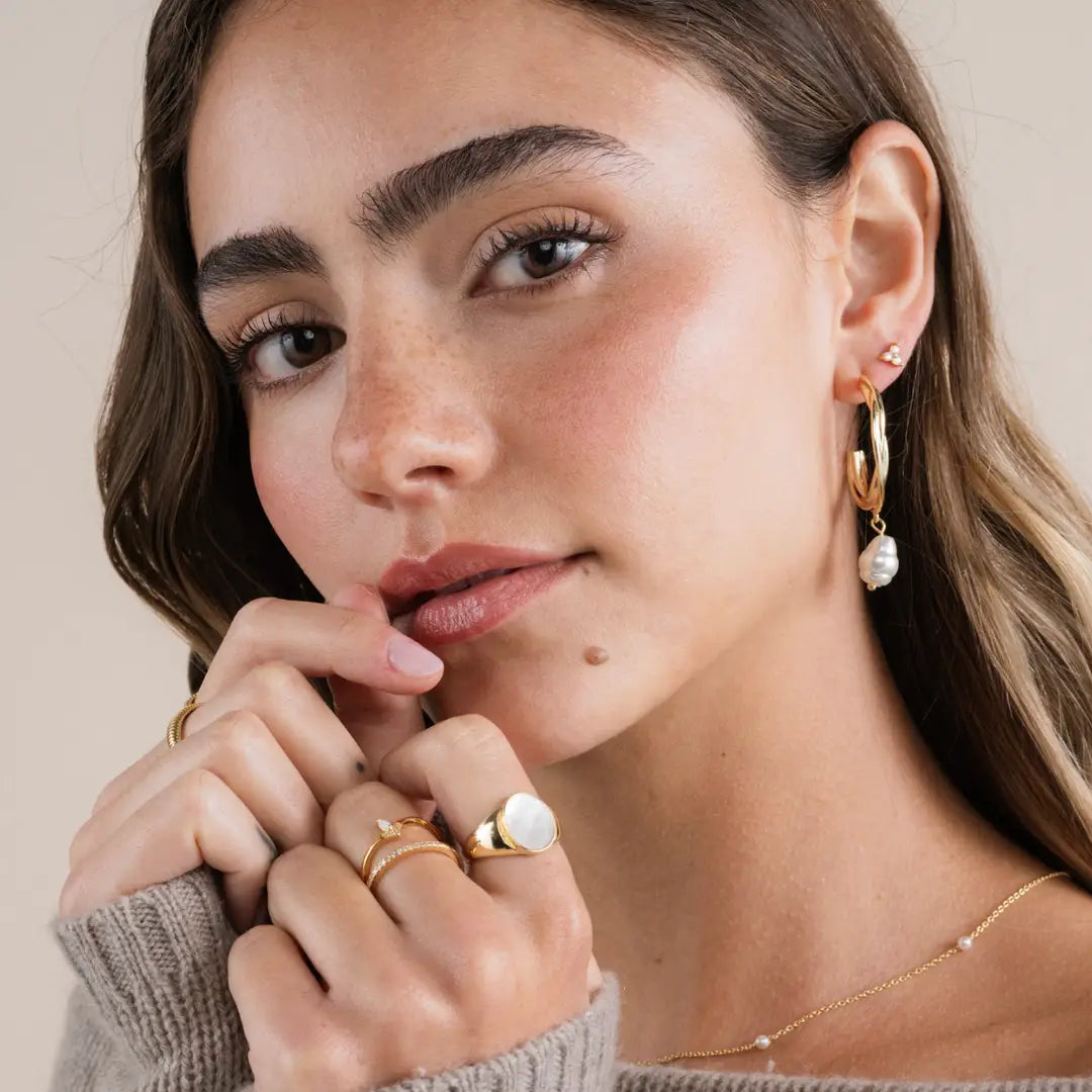 A young woman with long brown hair wears the JaxKelly Half Pavé Ring, gold hoop earrings with a white charm, and a necklace. She has natural makeup and gazes softly at the camera, gently touching her lips with her fingers.