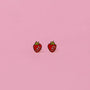 The Strawberry Enamel Flat Back Kids Earrings feature small, strawberry-shaped designs with red bodies, white seeds, and green tops—ideal for sensitive ears and shown side by side on a plain pink background.