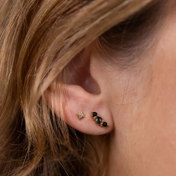 Close-up of a person's ear wearing JaxKelly Crescent Gold Earrings - Black Tourmaline: a star-shaped gold-plated stud and a curved bar with three black tourmaline stones on sterling silver posts. The person has light brown hair.