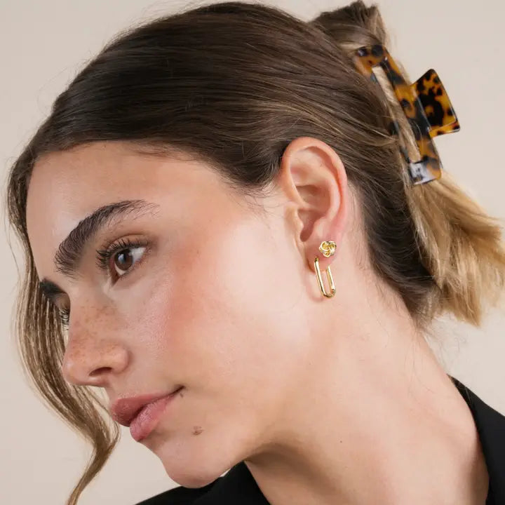 A woman with light skin and brown hair in a low bun, secured by a tortoiseshell clip, wears JaxKelly’s Petite Gold Knot Earrings. Featuring 18K gold plating, they add an elegant touch to her natural makeup and simple everyday style against a plain backdrop.