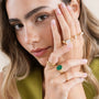 A woman with wavy light brown hair and natural makeup poses with her hands near her face, showcasing several gold rings including the JaxKelly Malachite Signet Ring. She wears a green top and has neatly manicured, light pink nails.