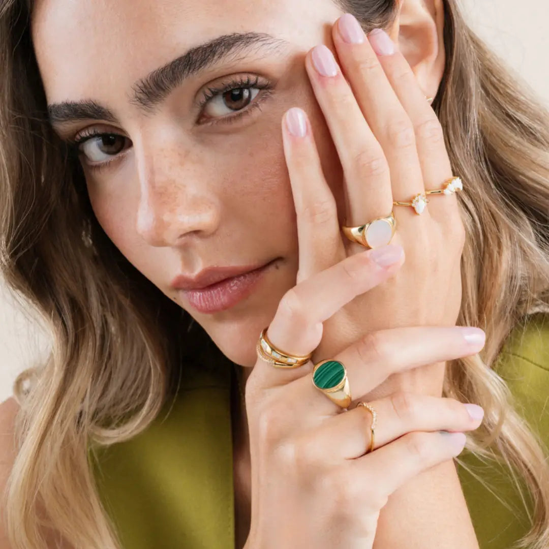 A woman with wavy light brown hair and natural makeup poses with her hands near her face, showcasing several gold rings including the JaxKelly Malachite Signet Ring. She wears a green top and has neatly manicured, light pink nails.
