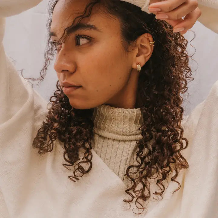 A person with curly hair wears a cream turtleneck and JaxKelly's Bold Hoop earrings—gold hoops with sterling silver posts—gazing sideways with one arm raised against a neutral background.