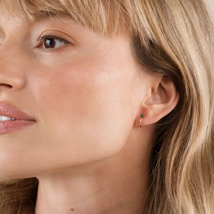 A close-up of a woman with light skin and blonde hair wearing JaxKelly's Open Mini Hoop Earrings – Blue Sapphire in her left ear, looking slightly to the left with a calm expression.