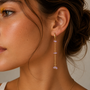 A close-up of a woman wearing the JaxKelly Raw Gemstone Chain Drop Earring - Lilac Quartz, featuring three lilac stones on a tarnish-free gold chain. The earring dangles by her ear as she sports smooth skin and natural makeup with yellow eyeshadow.