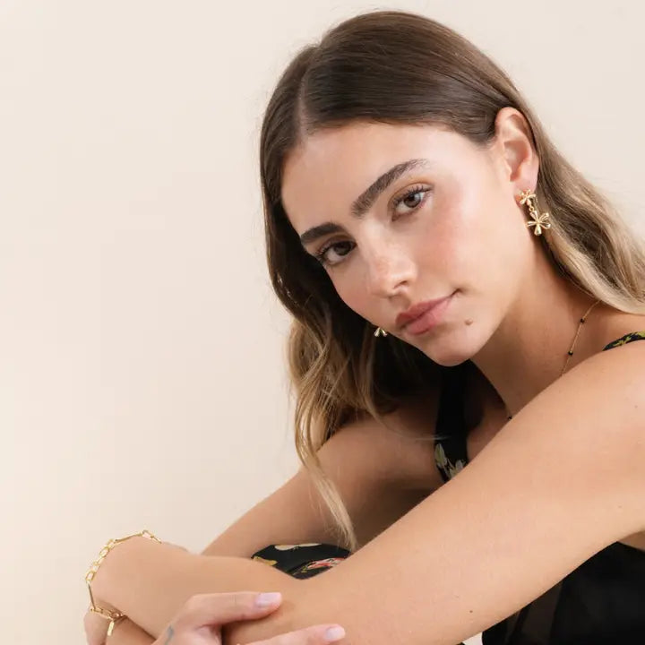 A woman with wavy brown hair and natural makeup sits with her arms on her knees, wearing JaxKelly's Double Daisy Drop Earrings, a gold bracelet, and a black floral top, set against a neutral background.