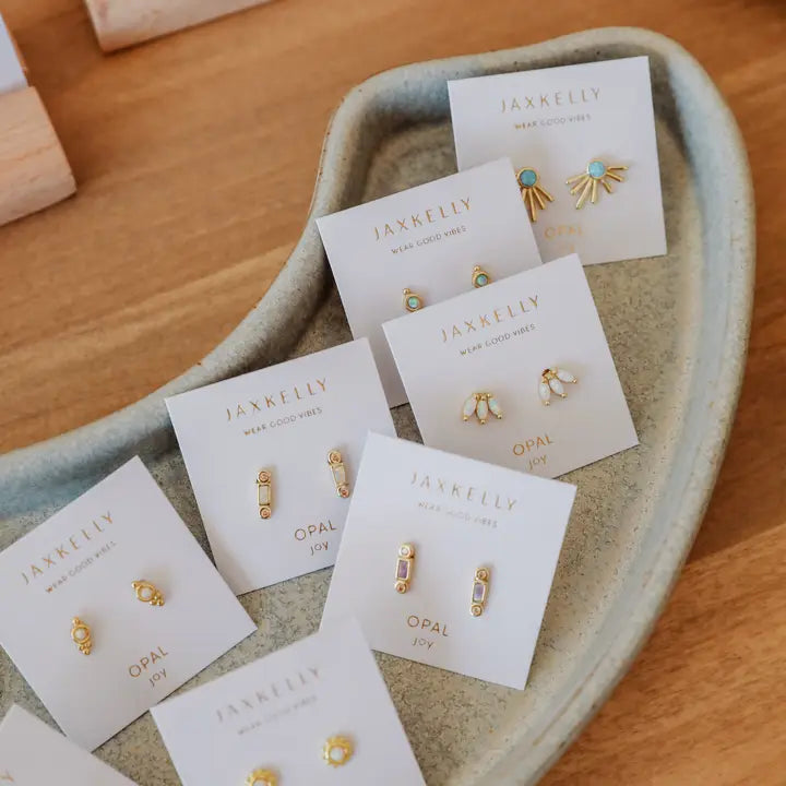 A display of JaxKelly White Opal Crown Stud earrings, showcased on branded white cards and arranged in a beige ceramic dish atop a wooden surface.