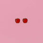 Two small, red Apple Enamel Flat Back Kids Earrings with green stems are side by side on a plain pink background—perfect hypoallergenic earrings for children.